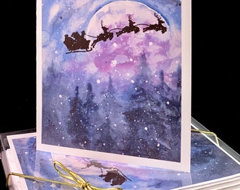 May include: A set of watercolor Christmas cards featuring a silhouette of Santa Claus flying in his sleigh pulled by reindeer in front of a full moon and a starry night sky.