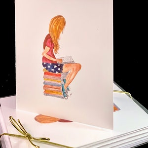 May include: A set of blank note cards with a watercolor illustration of a person sitting on a stack of books and reading. The person is wearing a red shirt, blue shorts with white stars, and blue sneakers. The note cards are tied together with a gold ribbon.