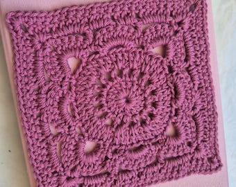 May include: A square, crocheted doily in a dusty rose color. The openwork design features a central floral motif surrounded by repeating patterns. The doily is placed on a light pink surface, showcasing the handmade texture.