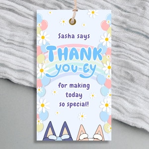 Może przedstawiać: Jasnoniebieska etykieta prezentowa z napisem "Sasha says THANK you-ey for making today so special!" w kolorowym wzorze. Etykieta jest ozdobiona stokrotkami, balonami i tęczą. U góry przymocowany jest sznurek.