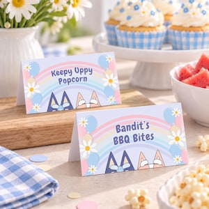 May include: Two rectangular folded paper food labels with a light blue background, a pastel rainbow, white daisies, and blue balloons. One label reads Keepy Uppy Popcorn and the other reads Bandit's BBQ Bites, both featuring cartoon dog ears at the bottom.