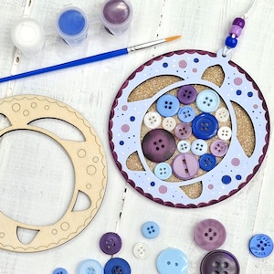 May include: A craft kit featuring a light blue and purple button-filled ornament with a cork center. Includes a wooden frame, paint pots, a paintbrush, and loose buttons in various shades of blue and purple. The ornament has a beaded string for hanging.