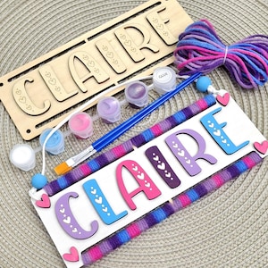 May include: A DIY craft kit featuring the name "CLAIRE" in wooden letters. The kit includes a wooden sign, paint pots in various colors, a paintbrush, glue, and colorful yarn. The letters are decorated with hearts, and the sign is ready for painting.