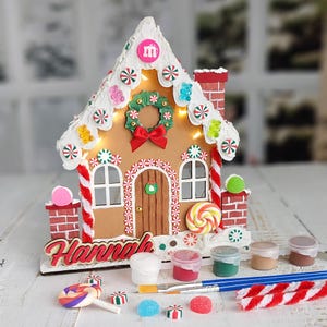 May include: A gingerbread house craft kit featuring a wooden structure with white icing, candy decorations, and a wreath. The name Hannah is displayed in red script. Includes paint pots, two paintbrushes, and candy-themed accessories for decoration.