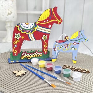 May include: A Dala horse craft kit featuring two wooden horses, one red and one blue. The kit includes paint pots, brushes, and the name "Jayden" on the base of the larger horse. The horses are decorated with stars.