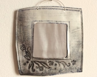 May include: A silver metal framed mirror with a decorative floral design around the edge. The mirror is hanging on a white wall.