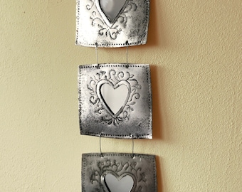 May include: Three silver metal wall hangings with heart-shaped cutouts and decorative scrollwork. Each square panel is connected by wire and hangs from a metal loop. The wall art has a distressed, vintage appearance.