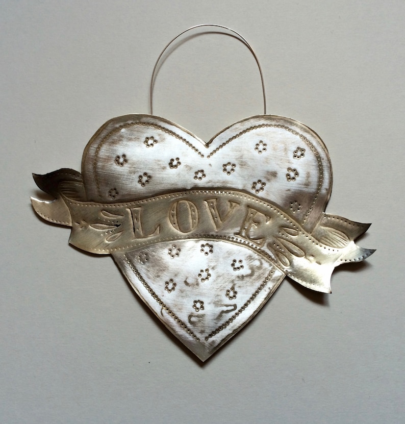 Puede incluir: Un adorno de metal blanco en forma de coraz&oacute;n con una pancarta que dice "LOVE" en estilo vintage. El coraz&oacute;n est&aacute; decorado con peque&ntilde;os dise&ntilde;os de flores en relieve.