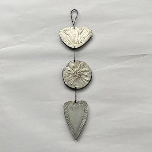 May include: A hanging decorative piece featuring three silver-colored, textured shapes: a petal-like top, a flower in the middle, and a heart at the bottom. Each shape is connected by a black string, creating a unique, handcrafted ornament.