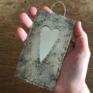 May include: A small, rectangular metal mirror with a heart-shaped cutout. The mirror has a distressed, aged finish and a white string hanger.