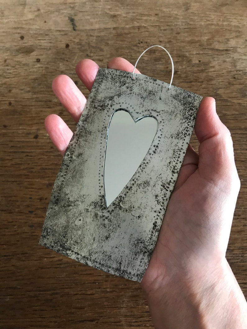 May include: A small, rectangular metal mirror with a heart-shaped cutout. The mirror has a distressed, aged finish and a white string hanger.