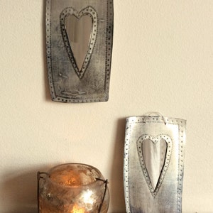 May include: Two silver metal mirrors with heart-shaped openings. The mirrors are hanging on a white wall. A candle holder with a lit candle is in the foreground.