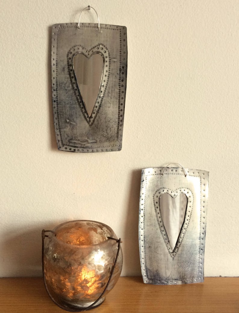 May include: Two silver metal mirrors with heart-shaped openings. The mirrors are hanging on a white wall. A candle holder with a lit candle is in the foreground.