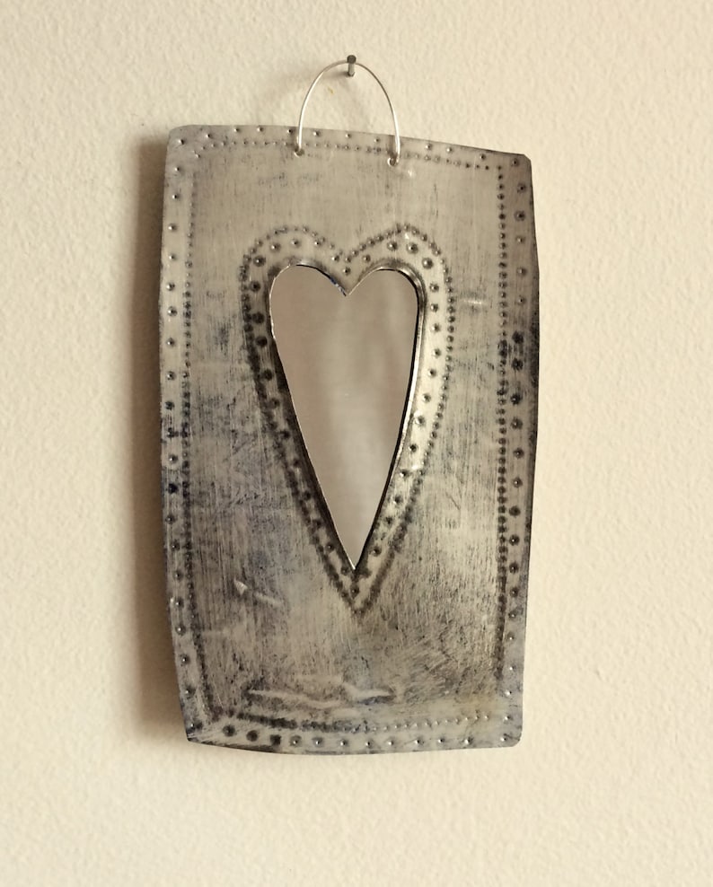 May include: A silver metal heart-shaped mirror with a distressed finish hangs on a wall. The mirror is surrounded by a decorative border of small metal dots.