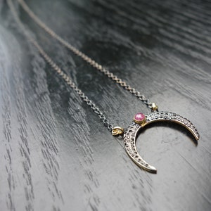 Pode incluir: Um colar com um pingente em forma de meia-lua e uma corrente prateada. O pingente tem uma moldura dourada, pedras pretas e uma pedra preciosa rosa no centro. O colar é exibido em uma superfície de madeira escura.