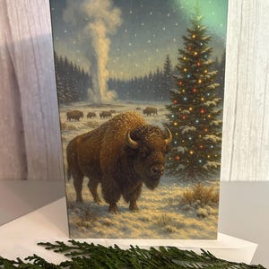 May include: A Christmas card featuring a winter scene with bison in a snowy landscape. A decorated Christmas tree stands to the right, and a geyser erupts in the background. The card is rectangular and has a glossy finish.