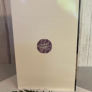 May include: A cream-colored greeting card with a purple circular sticker featuring the text "Paisley Prints '67" and a paisley design. The card is propped up against a wooden surface, with green foliage in the foreground.