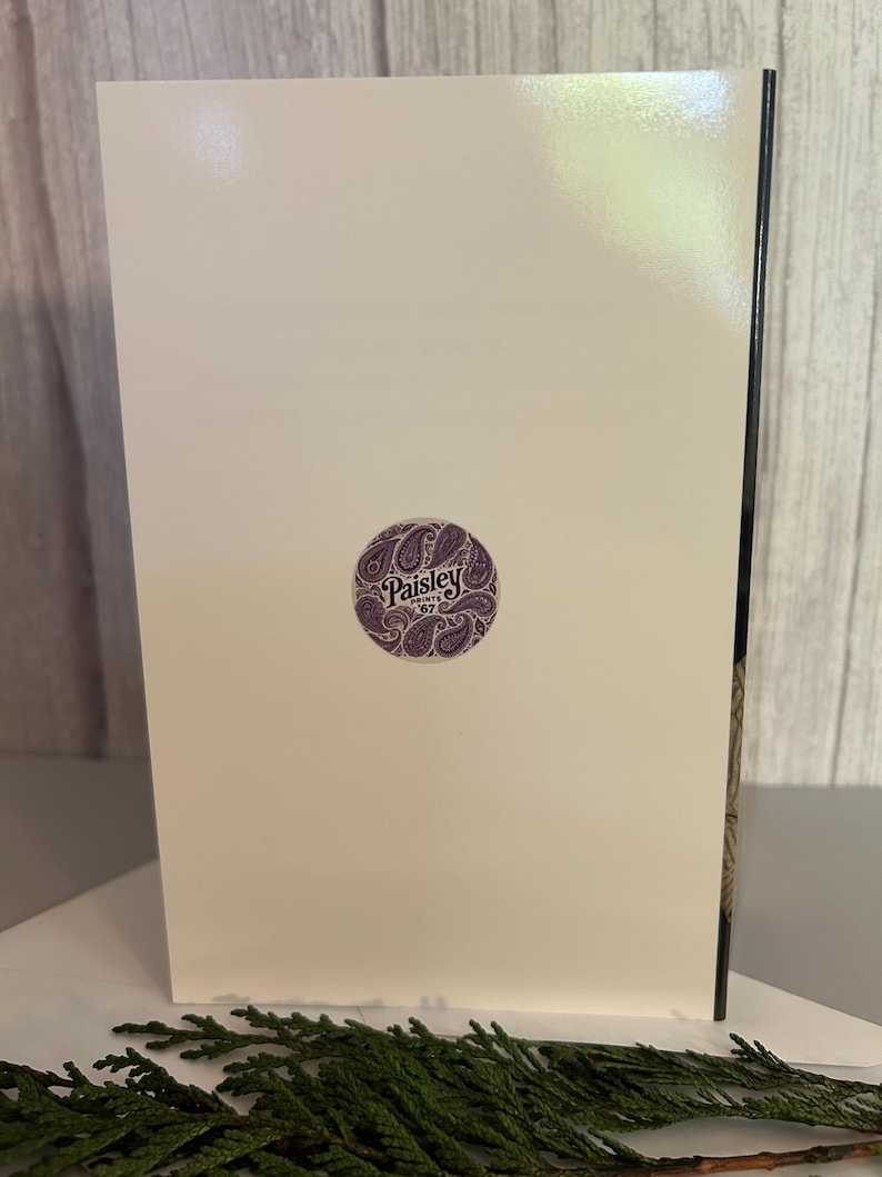May include: A cream-colored greeting card with a purple circular sticker featuring the text "Paisley Prints '67" and a paisley design. The card is propped up against a wooden surface, with green foliage in the foreground.