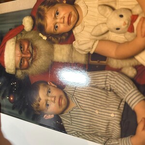 May include: A vintage photograph featuring Santa Claus with two children. The girl is holding a stuffed bunny with a red bow tie. The boy is wearing a striped shirt. All three subjects have a look of surprise on their faces.