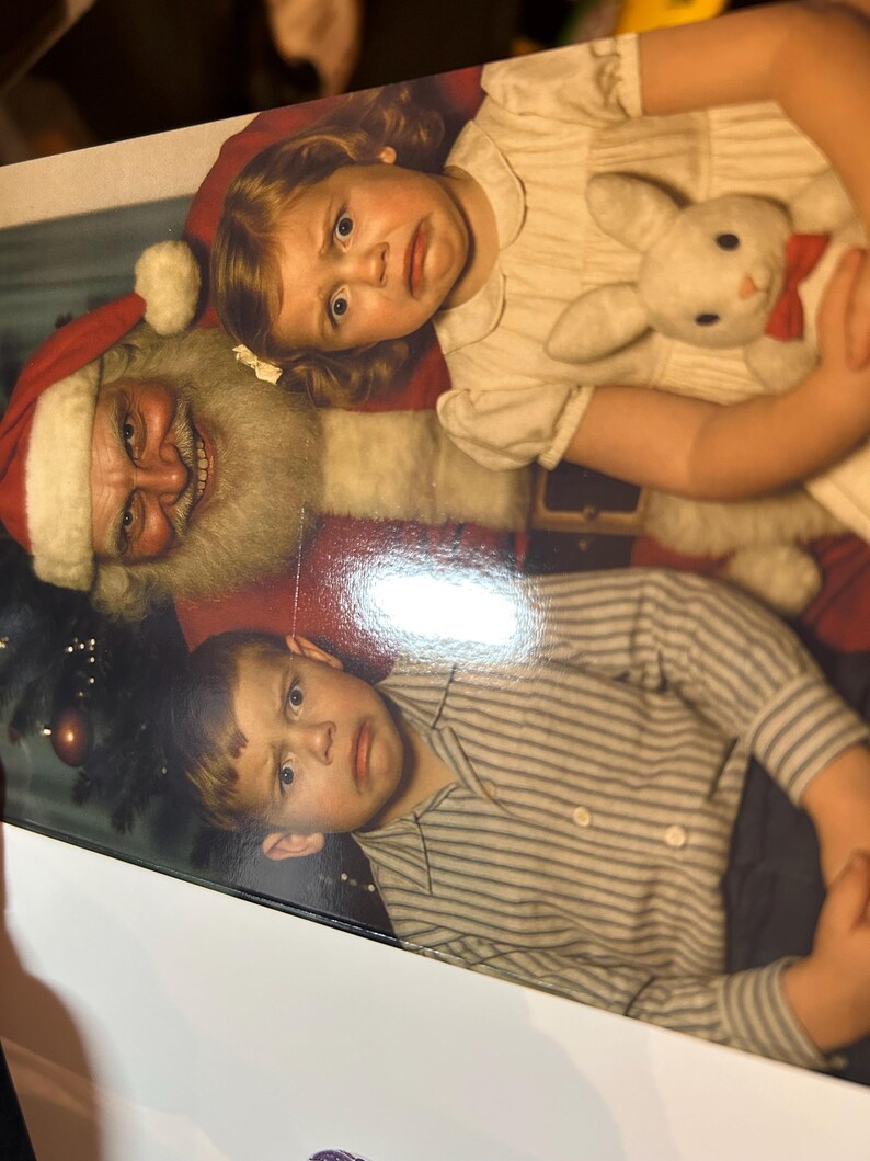 May include: A vintage photograph featuring Santa Claus with two children. The girl is holding a stuffed bunny with a red bow tie. The boy is wearing a striped shirt. All three subjects have a look of surprise on their faces.