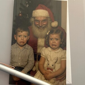 May include: A vintage Christmas card featuring Santa Claus with two children, all displaying expressions of fear. The children are dressed in a striped shirt and a white dress, and the girl is holding a stuffed bunny. Santa wears a red suit and hat.
