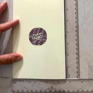 May include: A cream-colored greeting card with a circular purple sticker featuring a paisley design and the text "Paisley Prints '67". A ruler shows the card is approximately 4 inches wide.