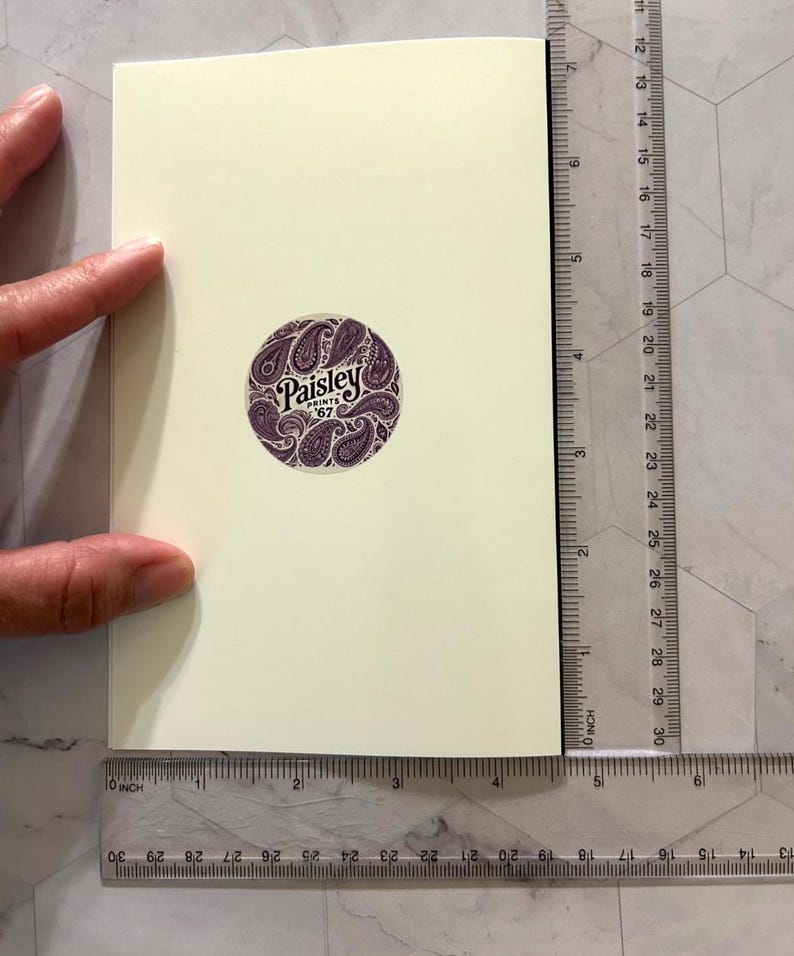 May include: A cream-colored greeting card with a circular purple sticker featuring a paisley design and the text "Paisley Prints '67". A ruler shows the card is approximately 4 inches wide.