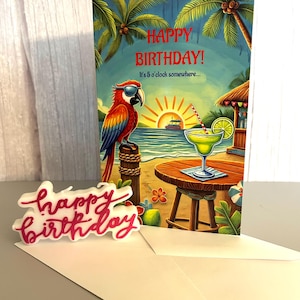 May include: A birthday card with a tropical theme. The card features a parrot wearing sunglasses, a margarita, and the text "Happy Birthday! It's 5 o'clock somewhere...". A separate "Happy Birthday" sign and an envelope are also present.