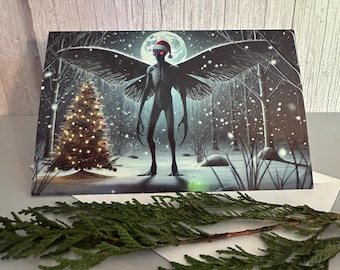 May include: A rectangular greeting card featuring a dark, winged creature wearing a red and white Santa hat, standing in a snowy forest at night with a glowing Christmas tree and a full moon in the background, placed near green pine needles.
