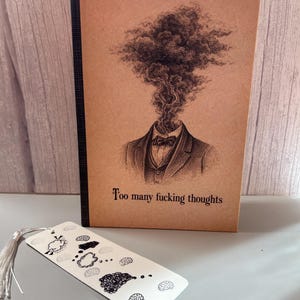 May include: A notebook with a brown cover featuring a detailed illustration of a person with smoke emanating from their head, and the text "Too many fucking thoughts." A white bookmark with brain illustrations and a silver tassel is in front.