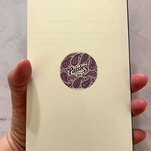 May include: A cream-colored notebook held open, revealing blank pages. A circular sticker with the text "Paisley Prints '67" in a decorative font is centered on the right page. The sticker features a paisley design in shades of purple.
