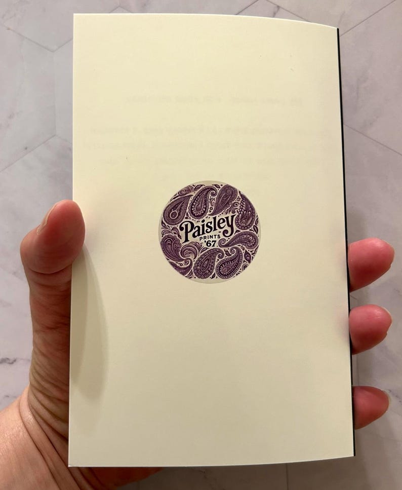 May include: A cream-colored notebook held open, revealing blank pages. A circular sticker with the text "Paisley Prints '67" in a decorative font is centered on the right page. The sticker features a paisley design in shades of purple.