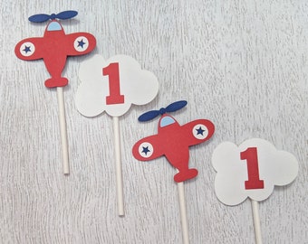 May include: Three red airplane cupcake toppers with blue propellers and white stars. Each airplane is paired with a white cloud with the number "1" in red.