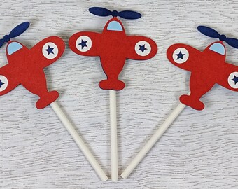 May include: Three red paper airplane cupcake toppers with blue propellers and white stars. The airplanes are attached to white sticks.