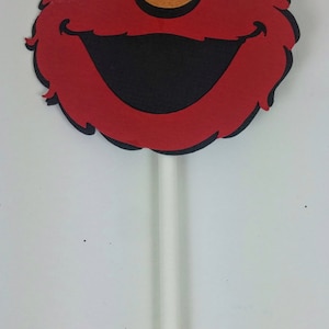 K&ouml;nnte beinhalten: Roter Filz-Elmo-Charakter-Gesichtsausschnitt auf einem wei&szlig;en Stiel. Der Charakter hat gro&szlig;e wei&szlig;e Augen, einen schwarzen Mund und eine rote Nase.