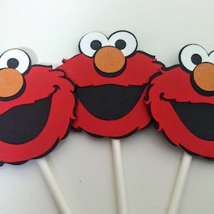 K&ouml;nnte beinhalten: Drei rote, flauschige Elmo-Cupcake-Topper aus Filz mit wei&szlig;en Augen, schwarzen M&uuml;ndern und orangenen Nasen. Jeder Topper ist an einem wei&szlig;en Stiel befestigt.