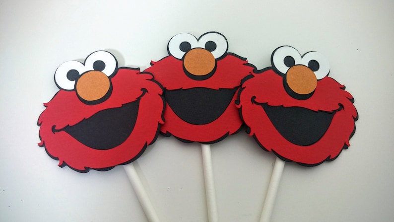 K&ouml;nnte beinhalten: Drei rote, flauschige Elmo-Cupcake-Topper aus Filz mit wei&szlig;en Augen, schwarzen M&uuml;ndern und orangenen Nasen. Jeder Topper ist an einem wei&szlig;en Stiel befestigt.