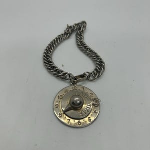 May include: A silver-toned charm bracelet with a circular pendant. The pendant features a clock face design with the words "DATE" and "TIME" and a central knob. The bracelet has a chain link design.