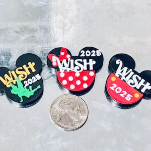 May include: Three black Mickey Mouse-shaped charms with the word "WISH" and the year "2025" in white. One charm has a green fairy, another has a red bow and polka dots, and the third has a red and yellow design. A quarter is next to the charms.