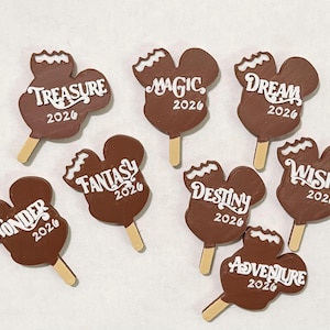 May include: A selection of brown Mickey Mouse-shaped ornaments resembling ice lollies, each with white text. The words are Treasure, Magic, Dream, Wonder, Fantasy, Destiny, Wish, and Adventure, with the year 2026.