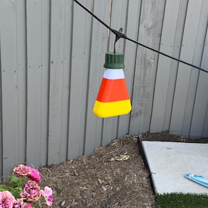 Könnte beinhalten: Eine Lichterkette mit einer candy-corn-förmigen Glühbirne mit horizontalen weißen, orangefarbenen und gelben Streifen und einer dunkelgrünen Kappe, die an einem schwarzen Kabel neben einer zweiten weißen Glühbirne mit dunkelgrüner Kappe hängt.