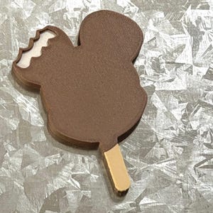 May include: A brown, mouse-shaped ice cream bar on a wooden stick. The top left ear of the chocolate-coated treat has a bite mark, revealing a white interior. The surface of the brown coating has a textured, matte finish, and the stick is light tan.
