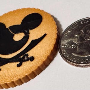 Könnte beinhalten: Ein beigefarbener, ovaler Spielchip mit einem schwarzen Piratenmotiv, das einen Mäusekopf und gekreuzte Schwerter zeigt. Eine US-Vierteldollarmünze liegt daneben zum Größenvergleich.