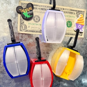 Puede incluir: Cuatro imanes de góndola en miniatura de colores azul, rojo, amarillo y lavanda. Cada uno tiene un asa negra y una puerta transparente segmentada. También se ve un billete de un dólar y dos imanes temáticos de Disney.