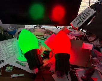 May include: Two large, teardrop-shaped light bulbs with black plastic bases. One bulb glows with a bright green light, while the other bulb glows with a vibrant red light. Both bulbs are positioned upright, emitting a strong, colorful illumination.