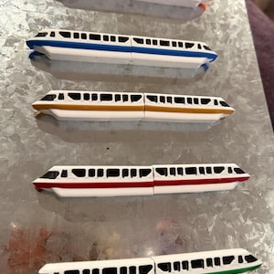 May include: Five toy monorail train cars in various colors: orange, blue, gold, red, and green. Each train car has a white body with black windows and colored accents along the top and bottom. The trains are arranged in a row.