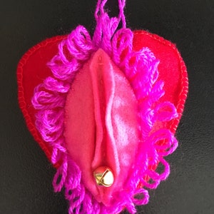 May include: A pink felt heart with a pink fringe border and a gold bell hanging from the bottom. The heart is attached to a pink string.