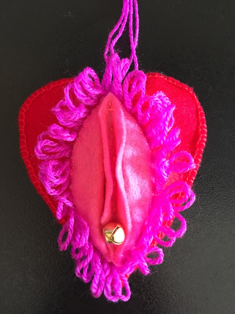 May include: A pink felt heart with a pink fringe border and a gold bell hanging from the bottom. The heart is attached to a pink string.
