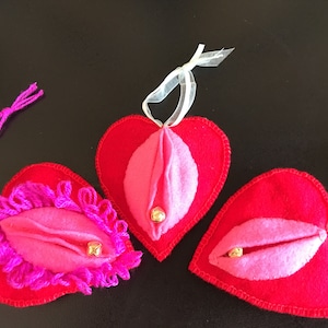 May include: Three red felt heart-shaped ornaments with pink felt interiors and gold bells. Each heart has a different design, one with pink yarn trim, one with a simple pink interior, and one with a more detailed pink interior.