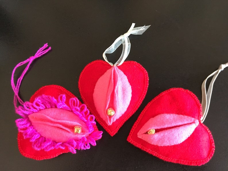 May include: Three red felt heart-shaped ornaments with pink felt interiors and gold bells. Each heart has a different design, one with pink yarn trim, one with a simple pink interior, and one with a more detailed pink interior.